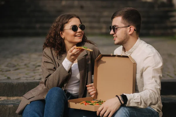 Young couple sitting on stairs outdoor and eating pizza. Handsome man with beaufitul woman taste pizza with vegetables. Vegan fast food