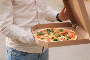 Açık havada pizza kutusunu açan genç bir adamın orta seçimi. Taze vejetaryen pizza, fesleğen, domates, brokoli ve laktozsuz peynir.