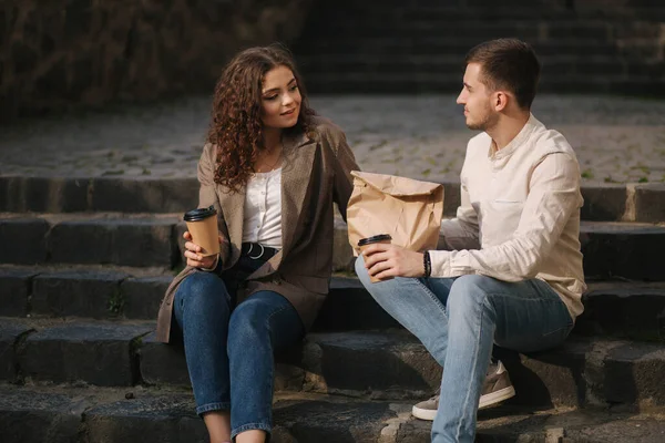 Pareja joven comida para llevar y sentarse al aire libre en las escaleras de la ciudad. Elegante pareja beber café y esperar amigos — Foto de Stock