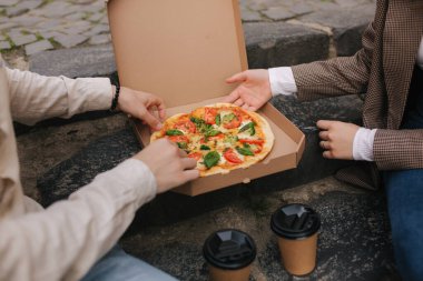Dışarıdaki kutudan birkaç dilim pizza kapıp yakın plan çekeceğiz. Erkek ve kadın elleri pizza yiyor. Taze domates fesleğenli vejetaryen pizza ve brokoli. Laktoz ve glütensiz.