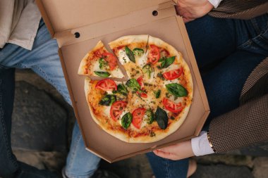 Dışarıdaki kutudan pizza alan çiftlerin en iyi görüntüsü. Erkek ve kadın elleri pizza yiyor. Taze domates fesleğenli vejetaryen pizza ve brokoli. Laktoz ve glütensiz. Pizza kutusu açık.