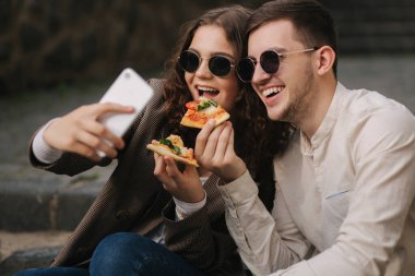 Genç blogcular pizza yerken selfie çekiyorlar. Güneş gözlüklü güzel çift kameralı telefona gülümsüyor. Vejetaryen fast food