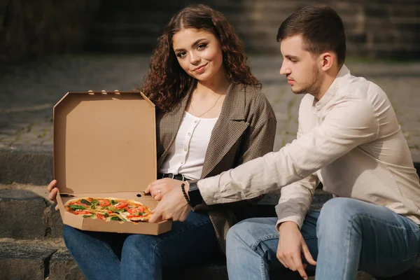 Ein junges Paar sitzt auf einer Treppe im Freien und isst Pizza. Schöner Mann mit Frau schmeckt Pizza mit Gemüse. Veganes Fast Food — Stockfoto