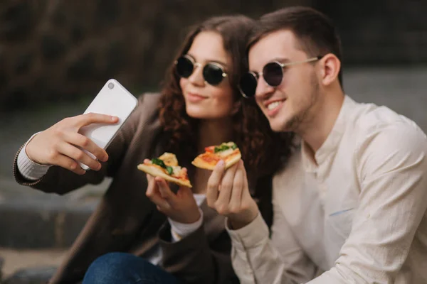 Νεαροί bloggers βγάζουν selfie τρώγοντας πίτσα. Όμορφο ζευγάρι με γυαλιά ηλίου χαμόγελο στην κάμερα του τηλεφώνου. Vegan fast food — Φωτογραφία Αρχείου