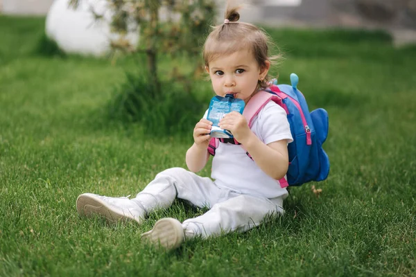 Imádnivaló kislány ül a füvön hátizsákkal. Aranyos kislány, gyümölcslevet iszik a szabadban. — Stock Fotó