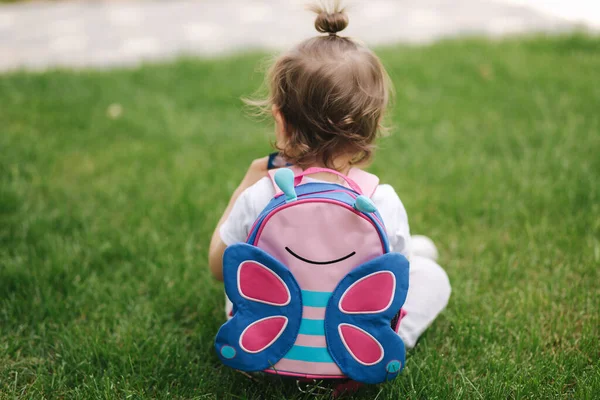 Vista posteriore della bambina con lo zaino si siede sull'erba all'aperto. spazio per il testo — Foto Stock