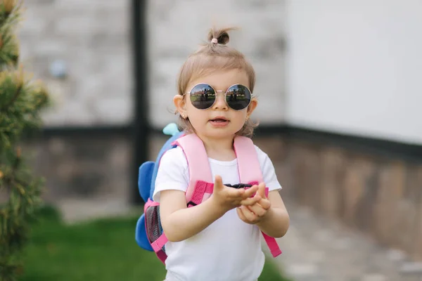 Portrait of Happy litle girl walk in gront of house with backpack. Cute one and half year girl in sunglasses — Stock Photo, Image