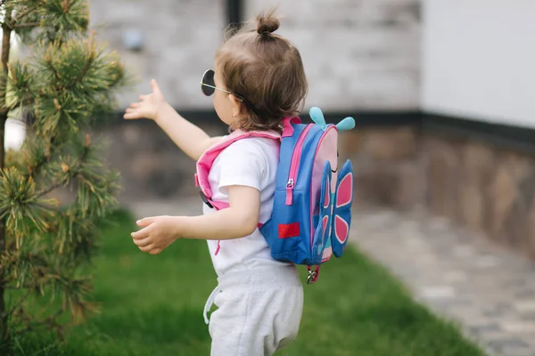 Portrét Happy Little Girl kráčí v grontu domu s batohem. Roztomilý jeden a půl roku dívka v slunečních brýlích — Stock fotografie