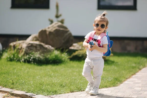 Šťastná holčička vejde do domu s batohem. Roztomilý jeden a půl roku dívka v slunečních brýlích — Stock fotografie