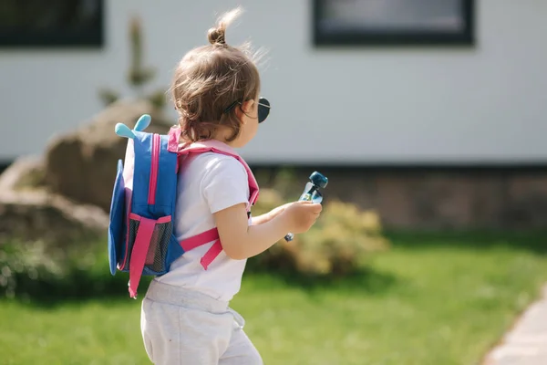 Happy litle girl cammina nel gront di casa con lo zaino. Carino un anno e mezzo ragazza in occhiali da sole — Foto Stock