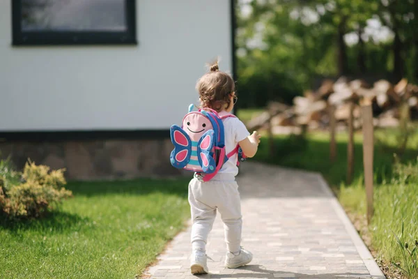 Happy litle girl cammina nel gront di casa con lo zaino. Carino un anno e mezzo ragazza in occhiali da sole — Foto Stock