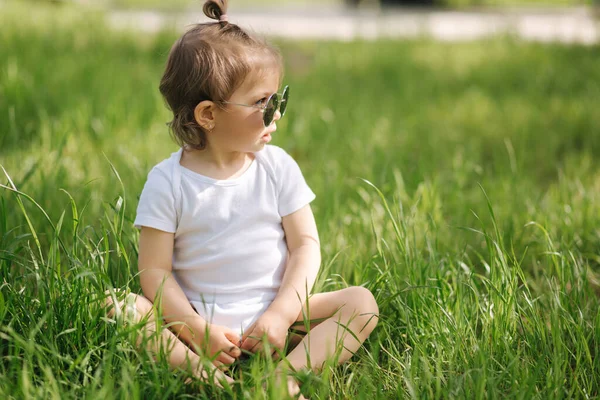 A boldog kislány a szabadban ül a fűben. Aranyos kislány fehér öltönyben és napszemüvegben az udvaron. Imádnivaló gyerek játszik a fűvel — Stock Fotó