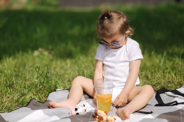 A boldog kisgyerek kint ül a szőnyegen és limonádét iszik. Imádnivaló kislány fehér ruhában. — Stock Fotó