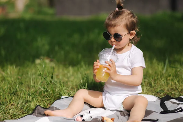 A boldog kisgyerek kint ül a szőnyegen és limonádét iszik. Imádnivaló kislány fehér ruhában. — Stock Fotó