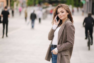 Kıvırcık saçlı çekici genç kadın şehirde telefon kullanıyor. Mutlu, güzel bir kadın telefonda konuşuyor.