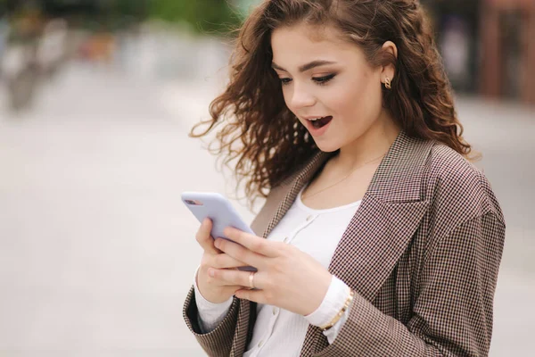 Молодая женщина с вьющимися волосами смотрит в телефон и удивляется. Счастливая женщина использовать телефон на открытом воздухе и поражен — стоковое фото