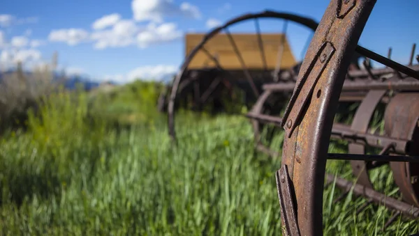 Farming ranch background with barn and rusty farm plow. Green grass, blue sky and wooden barn. Shallow depth of field with focus on rusty plow wheel. — Stock Photo, Image