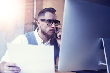 Sakallı işadamı Smartphone telefon görüştüğünüz kağıtları Hand.Young bulanık Desktop.Flare çalışan beyaz gömlek yelek giyen adam tutarak kullanarak başlangıç iş stratejisi Workplace.Man düşünüyorum.
