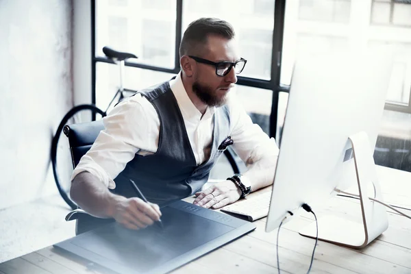 Diseño creativo barbudo Dibujo de trabajo Tableta digital Ordenador de sobremesa Mesa de madera.Joven con estilo con gafas Camisa blanca Chaleco Trabajo Moderno Loft Online Startup Project Borroso . — Foto de Stock