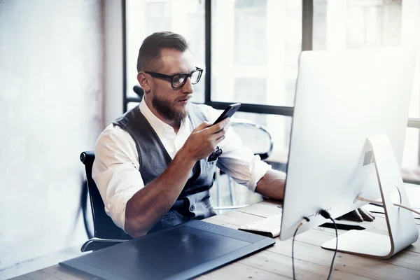 Bearded Stylish Young Man Wearded Glasses White Shirt Waistcoat Working Modern Loft Startup Process.Creative Person Using Smartphone Texting Message.Drawing Tablet Desktop Computer Wood Table.Blurred . — стоковое фото