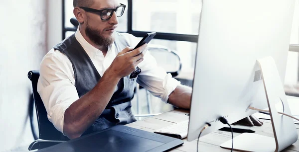 Bearded Stylish Young Man Wearing Glasses White Shirt Waistcoat Working Modern Loft Startup Process.Creative Person Using Mobile Phone Sending Message.Drawing Tablet Desktop Computer Wood Table.Wide.