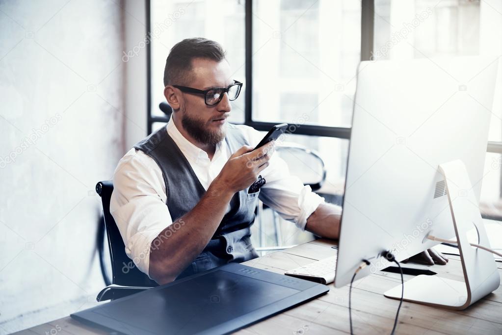 Bearded Stylish Young Man Wearing Glasses White Shirt Waistcoat Working Modern Loft Startup Process.Creative Person Using Smartphone Texting Message.Drawing Tablet Desktop Computer Wood Table.Blurred.