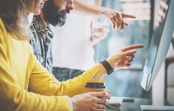 Closeup photo group of three young coworkers working together in a sunny office.Man typing on computer keyboard.Womans pointing hands to desktop screen.Horizontal image,blurred background.