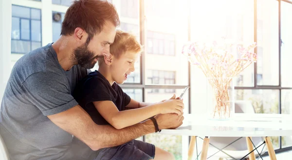 Young boy sitting with father at the table and playing together pc tablet in modern loft. Horizontal, blurred background. Sunlights effect. — Stock Photo, Image