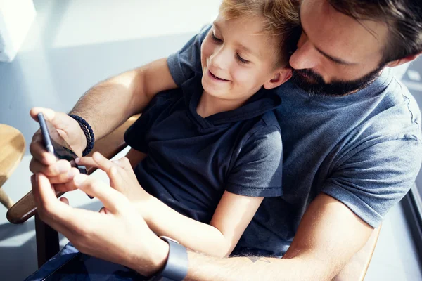 Zbliżenie młody chłopak siedzi z ojca i za pomocą smartfona w nowoczesne miejsce słoneczne. Poziome, niewyraźne tło. — Zdjęcie stockowe