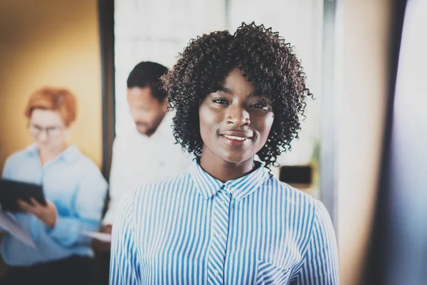 Ritratto di giovane donna africana con sguardo afro e sorriso alla camera.Business team sullo sfondo in ufficio moderno. Orizzontale, sfocato . — Foto Stock