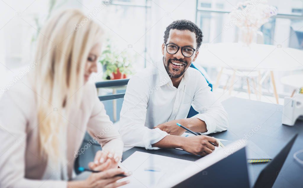 Two young coworkers working together in a modern office.Black man wearing glasses, looking at the camera and smiling.Woman discussing with colleague new project.Horizontal,blurred background