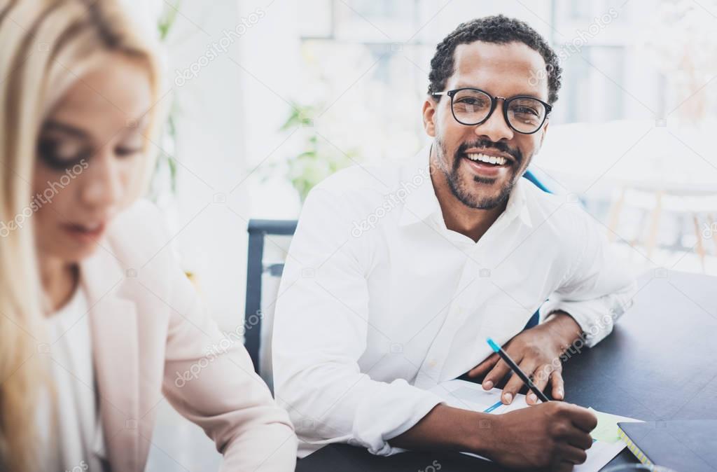 Two young business people working together in a modern office.Black man wearing glasses, looking at the camera and smiling.Businesswoman discussing with colleague new project.Horizontal,blurred .