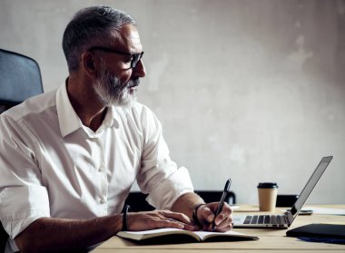 Klasik gözlüklü ve modern çatı katında ahşap masa ile dizüstü çalışan yetişkin başarılı iş adamı. Sakallı orta yaş erkek Notlar not defteri yapma. Yatay, bulanık arka plan.