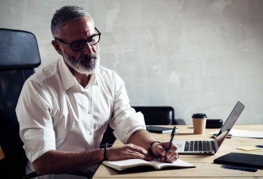 Klasik gözlüklü ve modern loft Studio ahşap masa çalışan yetişkin başarılı iş adamı. Orta yaş erkek Notlar not defteri yapma Beyaz gömlekli sakallı. Yatay, bulanık arka plan.