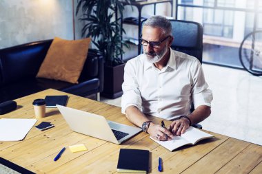 Klasik gözlüklü ve ahşap masa modern coworking ofiste çalışan yetişkin başarılı iş adamı. Şık sakallı orta yaş erkek Notlar not defteri yapma. Yatay, bulanık arka plan.