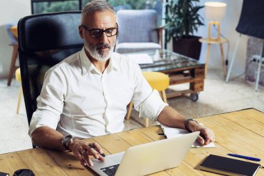 Klasik gözlüklü ve modern coworking Studio ahşap masa çalışan yetişkin başarılı iş adamı. Şık dizüstü bilgisayar üzerinde çalışma alanı kullanan orta çağı adam sakallı. Yatay, bulanık arka plan.
