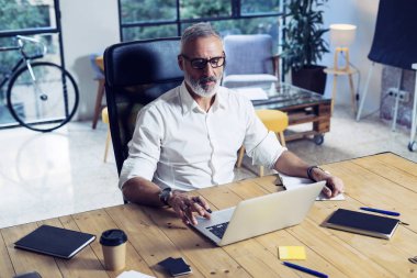 Klasik gözlüklü ve modern coworking Studio ahşap masa çalışan yetişkin başarılı iş adamı. Şık dizüstü bilgisayar kullanan orta çağı adam sakallı. Yatay, bulanık arka plan.
