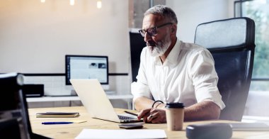 Klasik gözlüklü ve modern coworking Studio ahşap masa çalışan yetişkin profesyonel iş adamı. Şık dizüstü bilgisayar üzerinde çalışma alanı kullanan orta çağı adam sakallı. Yatay geniş, bulanık.