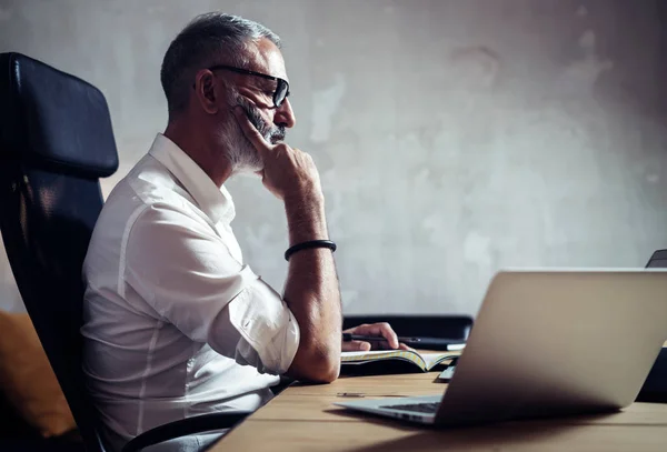 Homem de negócios barbudo adulto vestindo camisa branca e trabalhando na mesa de madeira no alto moderno.Homem fazendo anotações seu notebook.Horizontal, fundo borrado . — Fotografia de Stock