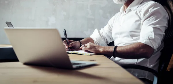 Erwachsene bärtige Geschäftsmann trägt eine klassische Brille und arbeitet am Holztisch in modernen Loft. Eleganter Mann macht Notizen in Notizbuch. horizontal breiter, unscharfer Hintergrund. — Stockfoto