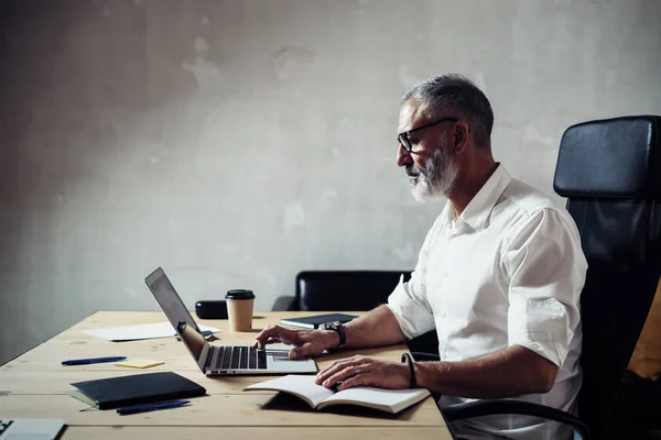 성인 수염된 사업가 클래식 안경 착용 하 고 현대 로프트에서 나무 테이블에서 일 노트북을 사용 하 여 세련 된 중 년 남자입니다. 수평, 흐린 배경. — 스톡 사진
