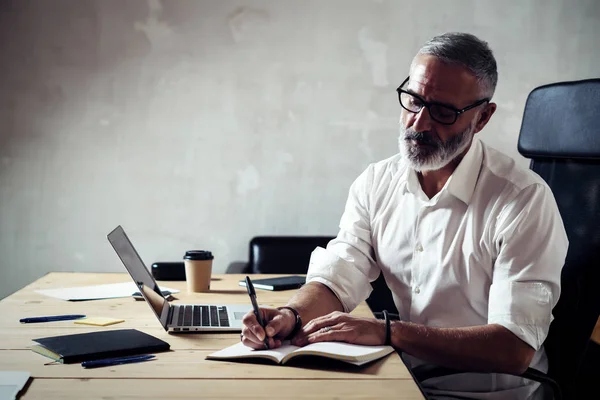 Adult stylish businessman wearing a classic glasses and working at the wood table in modern loft studio.Bearded middle age man in white shirt making notes notebook. Horizontal,blurred background. — Stock Photo, Image