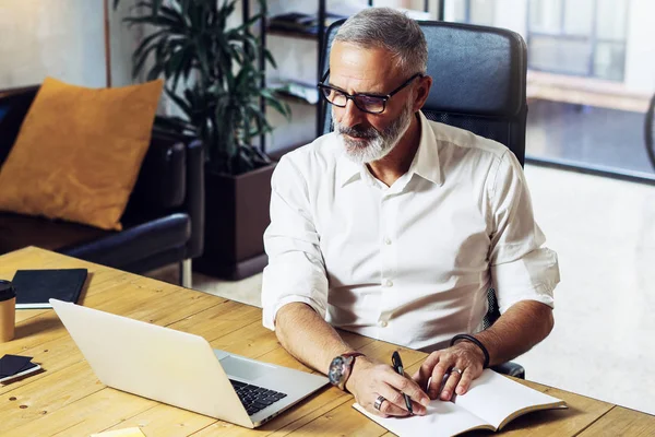 Adult successful businessman wearing a classic glasses and working at the wood table in modern coworking studio.Stylish bearded middle age man making notes notebook. Horizontal,blurred background. — Stock Photo, Image