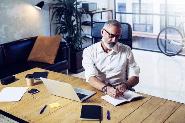 성인 성공적인 사업가 클래식 안경 착용 하 고 현대 coworking 스튜디오에서 나무 테이블에서 일 세련 된 수염된 중 년 남자 노트 노트북을 만들기입니다. 수평, 흐린 배경. — 스톡 사진