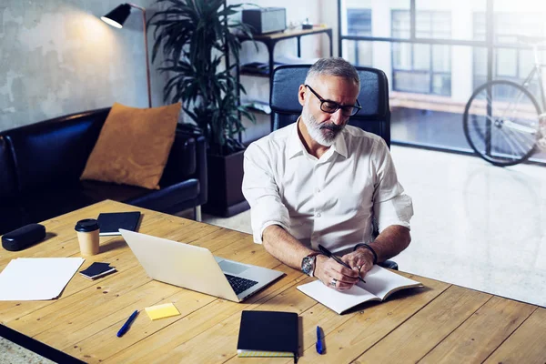 Adult successful businessman wearing a classic glasses and working at the wood table in modern coworking studio.Stylish bearded middle age man making notes notebook. Horizontal,blurred background. — Stock Photo, Image