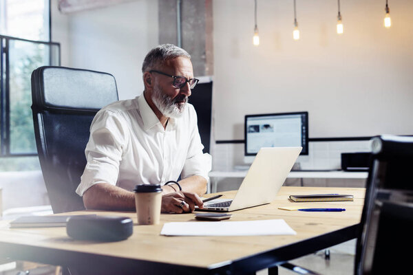 Adult professional businessman wearing a classic glasses and working at the wood table in modern coworking studio.Stylish bearded middle age man using laptop on workplace. Horizontal,blurred.