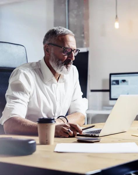 Adult professional businessman wearing a classic glasses and working at the wood table in modern coworking studio.Stylish bearded middle age man using laptop on workplace. Vertical,blurred. — Stock Photo, Image