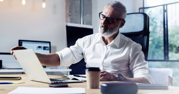 Middle age top manager wearing a classic glasses and working at the wood table in modern interior design office.Stylish bearded businessman using laptop on workplace. Horizontal wide,blurred.