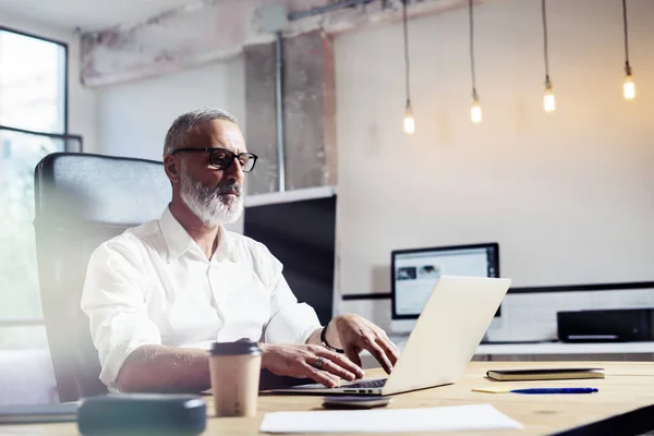 Finanzanalytiker mittleren Alters, der eine klassische Brille trägt und am Holztisch in einem modernen Einrichtungsbüro arbeitet.Stilvoller bärtiger Geschäftsmann mit Laptop am Arbeitsplatz. horizontal, verschwommen. — Stockfoto