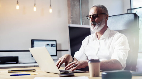 Middle age financial analyst wearing a classic glasses and working at the wood table with exchange markets.Stylish bearded businessman using laptop on workplace. Horizontal,blurred.